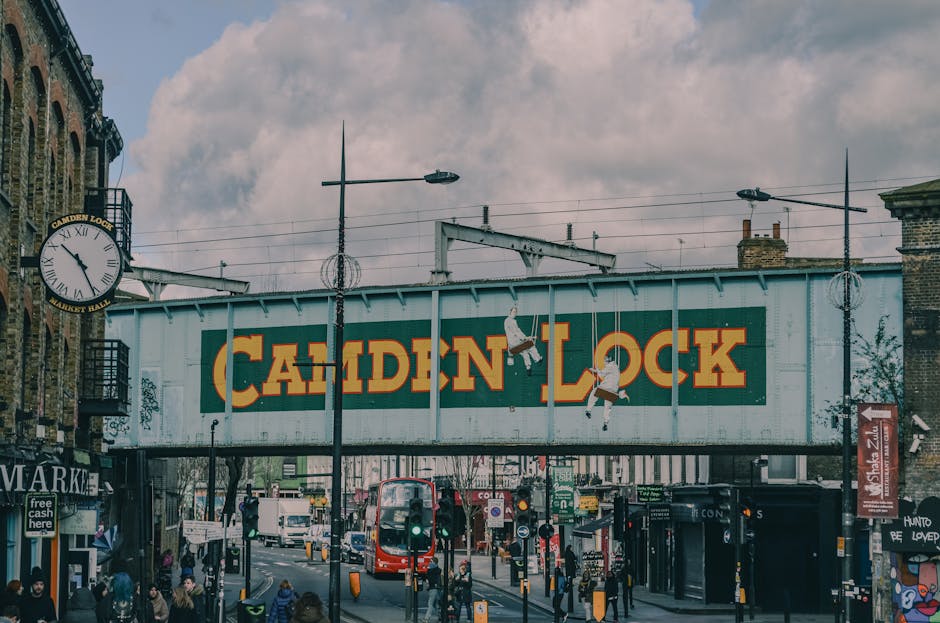 A street scene showing a large, painted metal bridge with the words 'Camden Lock' in bold yellow letters against a blue-green background, situated above busy pavement and road traffic in Camden, London. The bridge features a mural with two illustrated figures climbing and hanging from the structure. To the left, a traditional round clock with Roman numerals is mounted on a decorative metal bracket attached to a brick building. Below the bridge, pedestrians walk along the pavement, with some waiting at bus stops and others crossing the street. Several vehicles, including a red double-decker bus, are visible on the road, which is lined with shops and streetlights. The scene is illuminated by natural daylight with a partly cloudy sky above. The environment reflects a lively urban area typical of a busy part of Camden, ideal for showcasing moving or transport services offered by Lambeth Removals as part of home relocation or furniture transport in London.