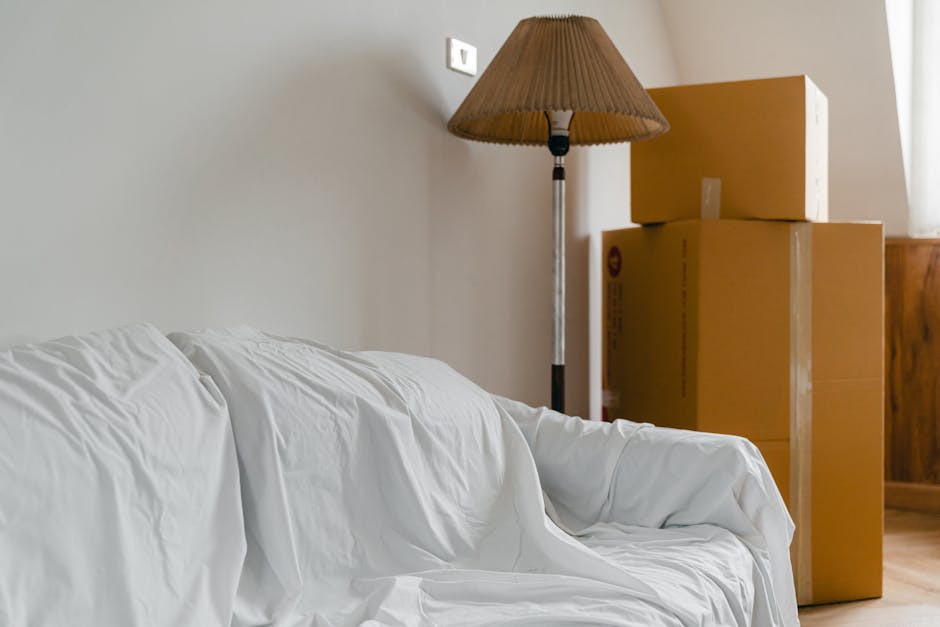 Inside a living room during a house relocation process, there are three large cardboard moving boxes stacked near a wooden cabinet against a beige wall. The boxes are sealed with clear packing tape, with one positioned on top of the other two. A tall floor lamp with a brown fabric lampshade stands next to the boxes, illuminating the area. In the foreground, a white fabric-covered sofa with visible creases is partially in view. The scene suggests preparation for furniture transport or packing and moving within a home, with natural light coming from a nearby window to the right, highlighting the packing materials placed ready for loading. Lambeth Removals, a professional moving service, is involved in this home relocation, evident from the organized placement of packing supplies and furniture, indicative of a systematic removal process.