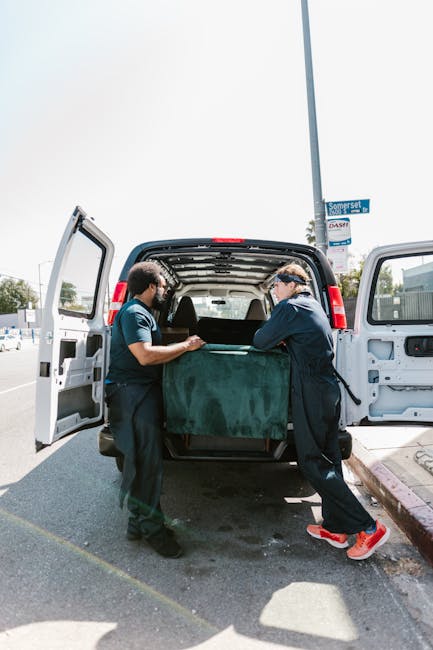 Two movers wearing dark uniforms and protective eyewear are loading a large, green padded furniture cover onto a black panel van equipped with sliding side doors in a parking area. The van's interior is empty, with visible wooden floors and a spacious cargo area suitable for home relocation. One mover is lifting the cover from the ground, while the other guides it into the vehicle, both focusing on careful handling of household furniture or boxes. The scene is set outdoors during daylight, with a street sign visible on a pole nearby, and in the background, a clear sky and some trees can be seen. This loading process reflects professional packing and moving procedures that Lambeth Removals might undertake during furniture transport and home relocations, emphasizing safe, efficient handling of belongings inside a vehicle designed for removals.