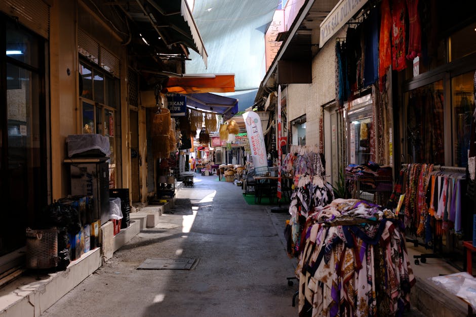 A narrow street during daytime lined with small shops and market stalls on both sides; on the right, a display of colourful scarves and fabrics drapes over tables, with some hanging on a rack, while on the left, shopfronts with glass windows and doors are visible, with outdoor items such as cardboard boxes, plastic containers, and small furniture pieces arranged along the pavement. Further along the street, more stalls are set up with hanging decorations, umbrellas, and signs, indicating ongoing market activity. The area is shaded by awnings and roof extensions, with natural light filtering through, creating highlights and shadows on the ground and shop displays. This scene captures the typical environment for home relocation or furniture transport services like those offered by Lambeth Removals, who may assist with packing, loading, and transporting household items through narrow streets such as this one in Brixton Market for clients undertaking interior moves or setting up new premises.