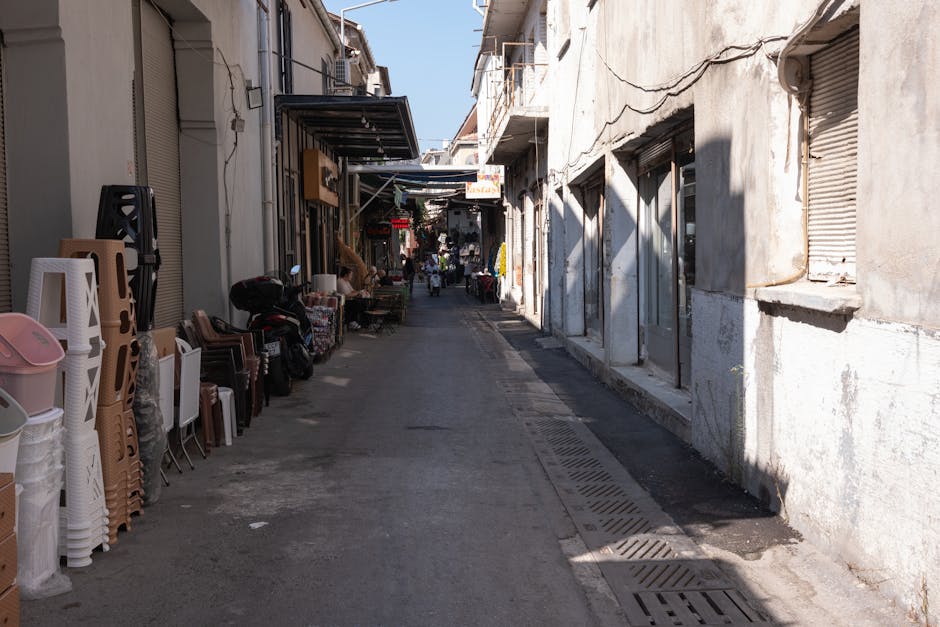 A narrow street in Brixton with commercial buildings on both sides, featuring closed shutters and an array of household furniture and boxes lined up along the pavement. On the left side, there are stacked plastic and wooden chairs, along with large cardboard boxes, plastic containers, and a black trolley partially visible. The right side shows a white building with closed metal shutters and a small window, with shadows cast on the wall from nearby structures. Several items are positioned near the entrances of shops, and the street appears to be used as a temporary storage or staging area during a home relocation process. In the distance, a few pedestrians walk further down the street under a clear sky, with overhead awnings providing some shade. The scene is well-lit, emphasizing the urban environment typical for moving and packing activities connected with house removals by Lambeth Removals, highlighting furniture transport, loading, and packing logistics for narrow street access during a house move.