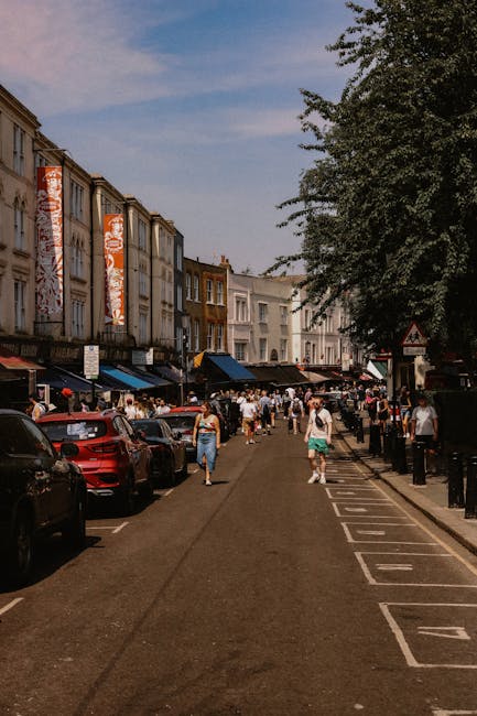 A narrow street during daytime lined with small shops and market stalls on both sides; on the right, a display of colourful scarves and fabrics drapes over tables, with some hanging on a rack, while on the left, shopfronts with glass windows and doors are visible, with outdoor items such as cardboard boxes, plastic containers, and small furniture pieces arranged along the pavement. Further along the street, more stalls are set up with hanging decorations, umbrellas, and signs, indicating ongoing market activity. The area is shaded by awnings and roof extensions, with natural light filtering through, creating highlights and shadows on the ground and shop displays. This scene captures the typical environment for home relocation or furniture transport services like those offered by Lambeth Removals, who may assist with packing, loading, and transporting household items through narrow streets such as this one in Brixton Market for clients undertaking interior moves or setting up new premises.