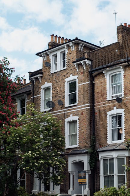This image depicts the exterior of a multi-storey brick residential building, characteristic of a typical London townhouse, with white-framed sash windows and decorative white detailing around the windows and roofline. The building's facade includes several satellite dishes attached to the brickwork. In the foreground, a flowering tree with green foliage partially obscures the lower part of the building, and a small sign indicating the presence of Lambeth Removals, a house removal and moving services company, is visible near the entrance. The sky is partly cloudy, and the building appears well-maintained, situated on a street where house moves, furniture transport, and packing logistics might be involved. The image suggests a location suitable for home relocation or furniture moving activities, with the building's accessible entryway and clear pathways for loaders and removal equipment.