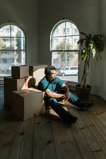 A man with curly hair, wearing a blue t-shirt and dark pants, sits on the wooden floor inside a room with large arched windows that allow natural light to fill the space. He is surrounded by several cardboard boxes secured with red packing tape, indicating an ongoing home relocation process. The boxes are stacked and positioned around him, some leaning against the wall, suggesting careful packing for a house move. In the background, the view through the windows reveals a street scene with parked cars and trees, typical of an urban environment. To the right, a tall potted plant with broad green leaves adds a touch of interior decor. The lighting creates a contrast between the brightly lit windows and the slightly shadowed interior, emphasizing the room’s spaciousness and the preparatory state of the moving process. Lambeth Removals specializes in local house and flat removals, supporting clients in packing and furniture transport, as reflected in the careful organization within this space.