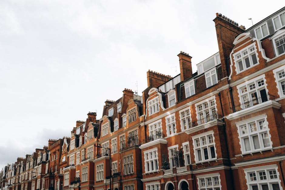The image shows a row of historic Victorian terraced houses in Kennington with red brick facades, white window frames, and decorative architectural details. Several windows are visible, some with small balconies or window boxes, and chimney stacks are prominent along the rooftops. The buildings are situated on a street with a cloudy sky overhead. In the foreground, a white moving van with open doors is parked on the pavement, and a few cardboard boxes and wrapped furniture items are being loaded into the vehicle by movers, evidencing a home relocation process. Protective blankets and straps are used to secure furniture, with the loading area adjacent to the entrance of a property. The scene reflects a professional move, coordinated by Lambeth Removals, with the process involving packing, furniture transport, and logistics typical of house removals in historic residential areas.