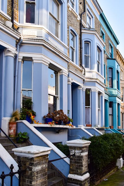 A row of Victorian terraced houses with ornate bay windows painted in various shades of blue, located in Kennington. The buildings feature decorative cornices and detailed brickwork, with small front gardens bordered by low brick walls and iron railings. The front steps, leading up to the entrances, are visible, with some plants and flowers placed in pots near the doorways. The pavement and stairs are in the foreground, with a black iron gate and brick pillars at the staircase entrance. Bright daylight illuminates the scene, highlighting the architectural details. This image captures the typical residential environment suitable for house removals or furniture transport services by Lambeth Removals, emphasizing careful packing and loading activities associated with relocating homes in Victorian terraces and estate properties in Lambeth. The setting provides context for professional moving or home relocation logistics in historic residential areas.