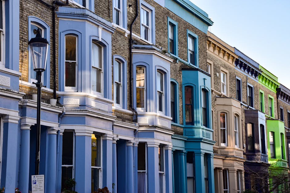 The image shows a row of historic Victorian terraced houses in Kennington with red brick facades, white window frames, and decorative architectural details. Several windows are visible, some with small balconies or window boxes, and chimney stacks are prominent along the rooftops. The buildings are situated on a street with a cloudy sky overhead. In the foreground, a white moving van with open doors is parked on the pavement, and a few cardboard boxes and wrapped furniture items are being loaded into the vehicle by movers, evidencing a home relocation process. Protective blankets and straps are used to secure furniture, with the loading area adjacent to the entrance of a property. The scene reflects a professional move, coordinated by Lambeth Removals, with the process involving packing, furniture transport, and logistics typical of house removals in historic residential areas.