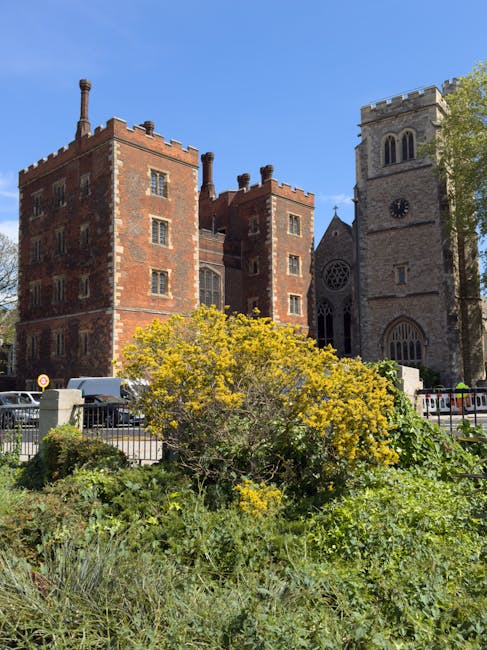 A street scene in front of a historic red brick castle-like building with multiple turrets and chimneys, situated behind a small garden area with green shrubs and a flowering yellow bush. The garden is enclosed by a metal fence, and behind it, several parked cars are visible along the street. To the right, part of a stone church with a high, square clock tower and arched windows can be seen, set against a bright blue sky with some trees nearby. The setting suggests a residential or mixed-use area where house removals or moving services by Lambeth Removals may operate, with a focus on the external environment and surrounding architecture during a home relocation process.