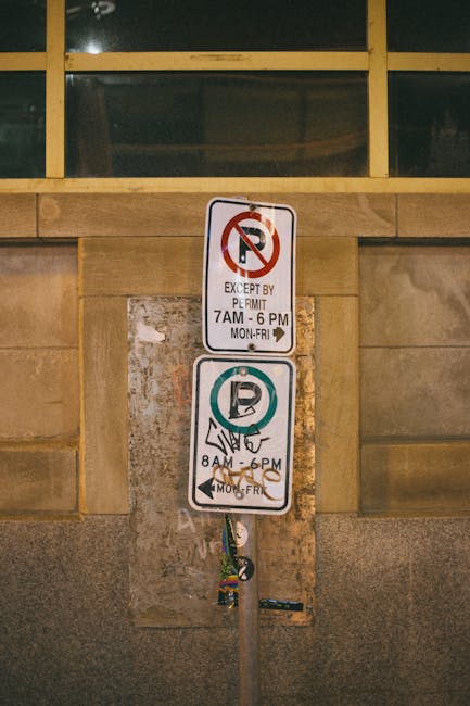 A metal parking restriction sign mounted on a pole is positioned against a stone wall beneath a row of high windows with yellow frames. The sign displays two sections: the top section indicates no parking except by permit from 7 am to 6 pm, Monday through Friday, with a red circle and slash over a letter 'P'. The lower section shows a permit parking symbol with a green circle and a white 'P' inside, and notes permit only parking from 8 am to 6 pm, Monday through Friday, also with a visual depiction of a permit sticker being displayed. The sign is part of a street scene related to house removals and moving logistics, where Lambeth Removals may need to navigate parking rules during furniture transport or home relocation activities. The environment suggests an urban setting with focused lighting and the sign serving as a guide for parking compliance during loading or unloading procedures associated with house moves.