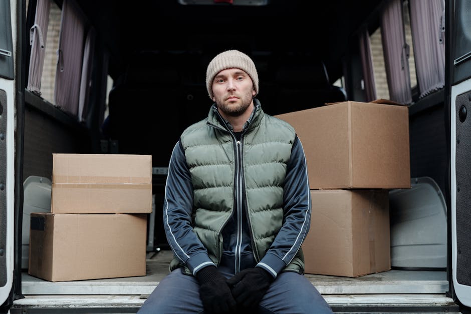 A man with a serious expression wearing a grey knitted beanie, a green puffer vest, navy blue track jacket, and black gloves, sitting on the edge of an open van loaded with several brown cardboard moving boxes of various sizes, some sealed with packing tape, inside a residential driveway or street. The van's interior is visible, showing a dark cargo space, and the boxes are arranged with some on the floor and others stacked. The environment includes a paved surface and part of a house or building in the background, with natural daylight illuminating the scene, indicative of a house relocation or furniture transport process. Lambeth Removals specializes in efficient, same-day house removals, and this loading process is part of their professional moving services.