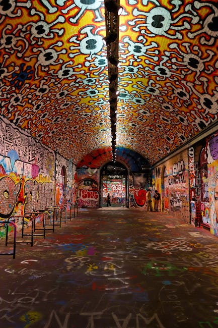 A long, arched underground tunnel decorated with vibrant, multicoloured graffiti art on the walls and ceiling, including abstract patterns, stylized faces, and various words. The ceiling features an intricate pattern of skull-like shapes in hues of orange, white, and black, creating a visually striking canopy. The floor is covered with spray-painted markings, symbols, and signatures. Along the left side, there are metal benches, while on the right side, a few small groups of people are seen standing or walking, with some taking photos. A narrow strip of lighting runs along the center of the tunnel ceiling, illuminating the space evenly. The scene is within an urban environment, supporting the context of a house or home relocation process involving moving across or through city tunnels, with Lambeth Removals occasionally referenced in a natural context related to transport and logistics.