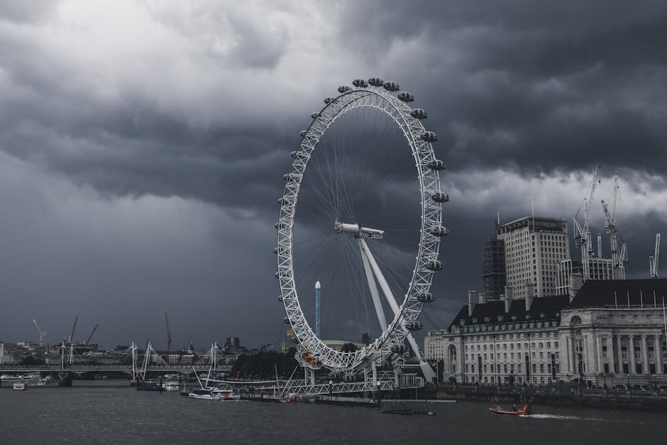 A large Ferris wheel situated on the bank of a river in an urban setting, under a dark, cloudy sky. The wheel has multiple enclosed gondolas attached around its circumference and is partially illuminated, with some gondolas visible against the overcast sky. The surrounding area includes tall buildings, some under construction with cranes visible, and a bridge spanning the river. The scene appears to be taken during stormy weather, with the sky heavy with dark clouds, creating a dramatic atmosphere. Though not directly related to house removals, this image could metaphorically represent the complexity of home relocation or transport logistics, as visualized by the towering Ferris wheel in the cityscape, with Lambeth Removals providing professional moving services.