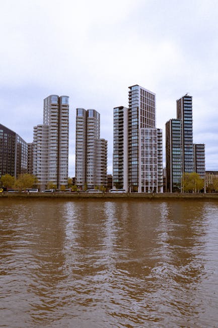 A view of modern high-rise buildings along the riverbank, with the buildings constructed mainly from glass and concrete, reflecting the overcast sky. The buildings are part of an office or residential development visible from across the water, with some featuring rounded and rectangular shapes. In the foreground, the river has a brownish hue with slight ripples, indicating gentle water movement. The scene appears to be outdoors during daytime with natural lighting. This image relates to house and office removals, illustrating the urban environment where furniture, boxes, and equipment might be transported during a relocation process, as managed by companies like Lambeth Removals at Vauxhall SE11 near Nine Elms, focusing on moving logistics, packing, and furniture transport in city settings.
