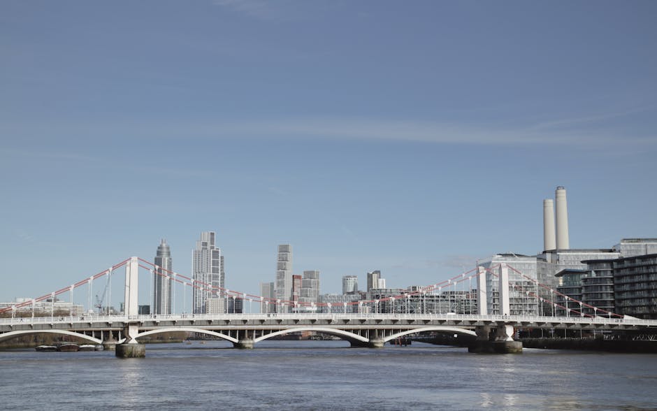 A view of the London skyline featuring several modern office buildings with reflective glass exteriors, situated along the River Thames. In the foreground, a boat is moored near the riverbank, with the water displaying gentle ripples. On the shore, there are some low-rise structures and a partially constructed or renovated building with visible structural elements. The sky above shows scattered clouds and natural daylight, creating a clear visibility of the high-rise commercial architecture. This scene relates to urban infrastructure and business districts, which are often involved in city-based removals and relocation services, supported by companies like Lambeth Removals specializing in house and office removals near central London locations such as Vauxhall SE11 near Nine Elms.