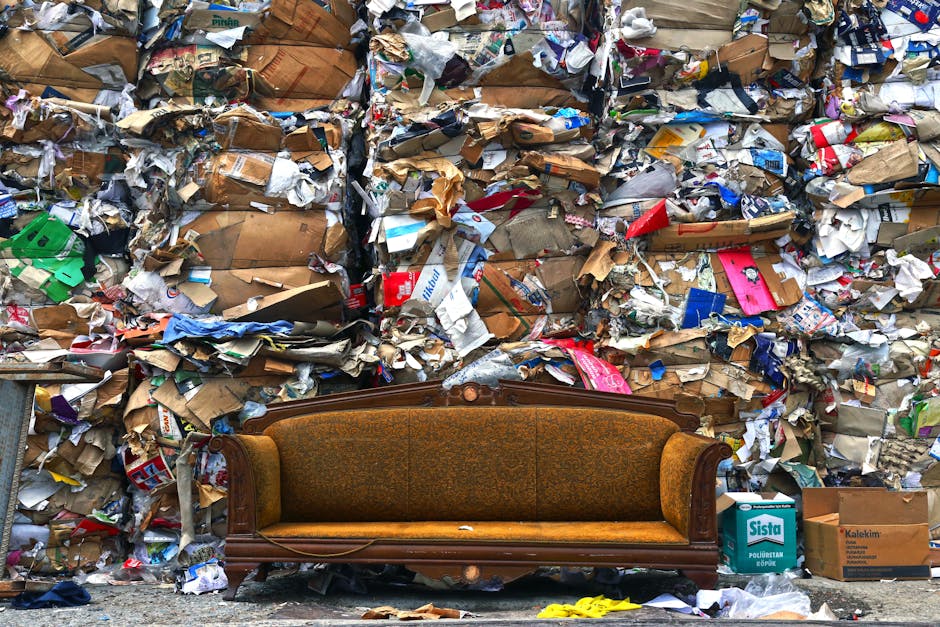 An outdoor scene showing a vintage wooden sofa with brown patterned upholstery positioned on a pavement in front of a large wall covered with a dense accumulation of mixed waste, including crumpled cardboard boxes, plastic packaging, paper waste, and discarded packaging materials. The sofa appears old, with some visible wear and tear, and is situated close to the waste debris. Surrounding the sofa are additional cardboard boxes and plastic bags, with some items spilling onto the ground, indicating improper waste disposal. This scene depicts a situation where furniture or household waste has been dumped improperly, illustrating the importance of following waste disposal rules in Lambeth for furniture removals, as promoted by Lambeth Removals, a professional removals service. The environment is lit naturally, highlighting the cluttered and untidy state, emphasizing the need for proper waste management during home relocation or furniture transport.