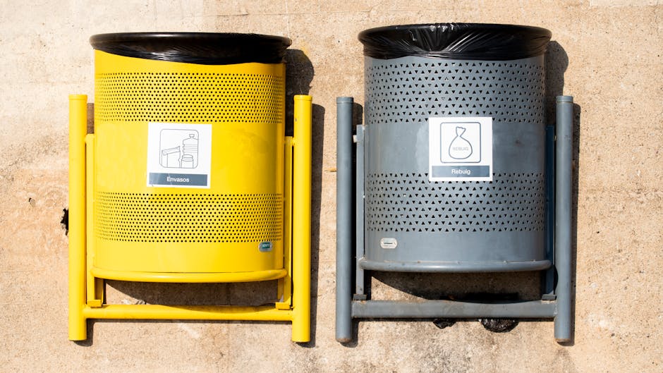 Two cylindrical waste collection bins placed side by side on a paved surface outside a property, with sunlight casting shadows. The left bin is yellow with a black lid, featuring a label indicating it is for packaging waste, and has small perforations around its middle section. The right bin is grey with a black lid, displaying a label for reusable materials, with a pattern of triangular cutouts around its middle and a different label illustration. Both bins are supported by metal frames, with the yellow bin's frame painted yellow and the grey bin's frame painted grey. The footwear and belongings of residents are not visible, and the environment suggests a typical residential area prepared for waste disposal during a home relocation or furniture transport process. Lambeth Removals provides relevant house removal services, including proper waste disposal following local regulations concerning furniture and packaging materials during moves in Lambeth.